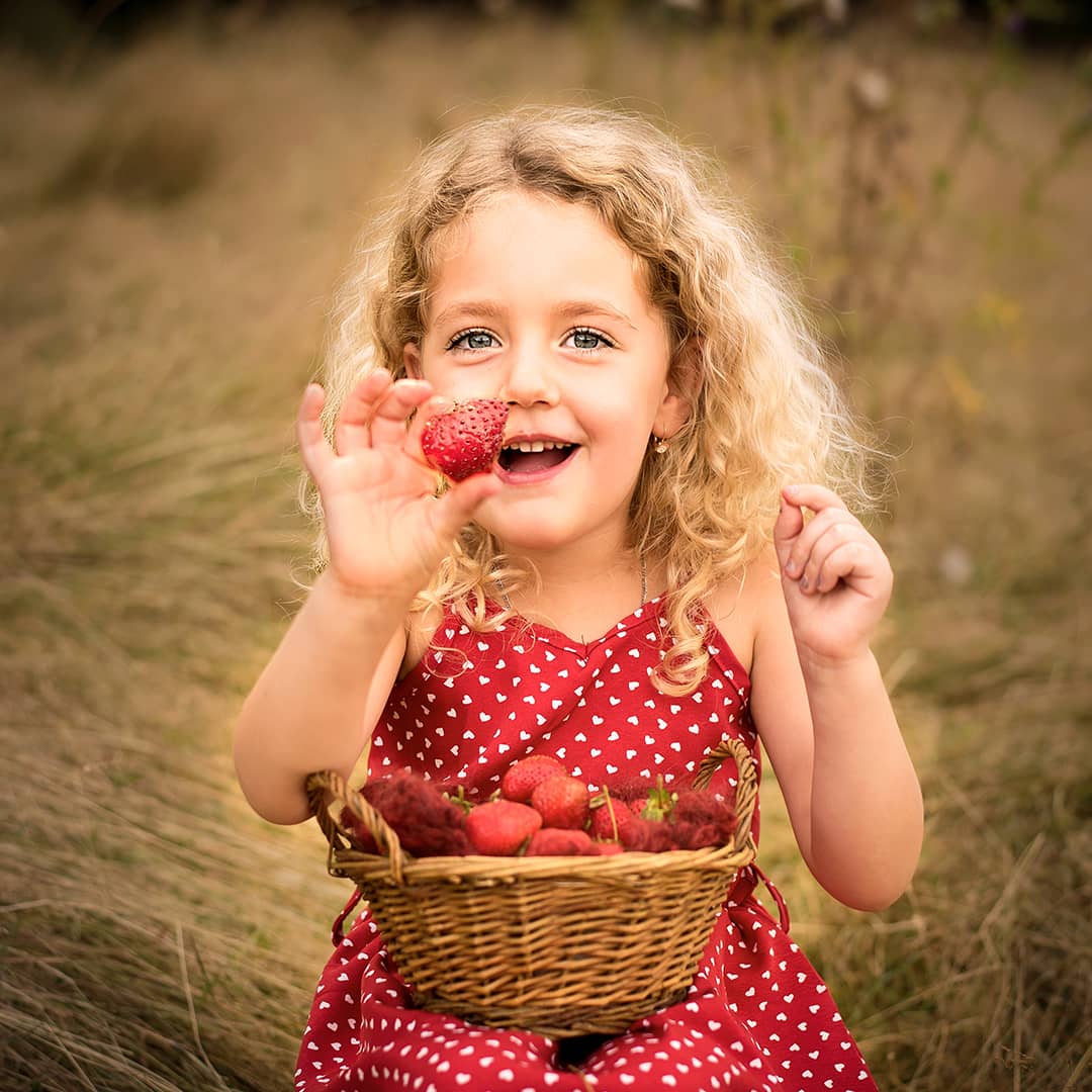 Fröhliches blondes Kind zeigt stolz eine Erdbeere in die Kamera, während es mit einem Weidenkorb im Sommer-Gras sitzt beim Kinderfotoshooting in Wuppertal