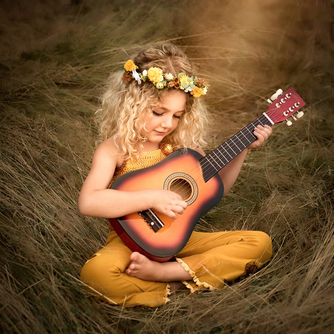 Glückliches Kind mit blonden Locken und Blumenkranz spielt im Gras auf einer kleinen Gitarre beim ungestellten Erlebnis-Fotoshooting im Raum Wuppertal