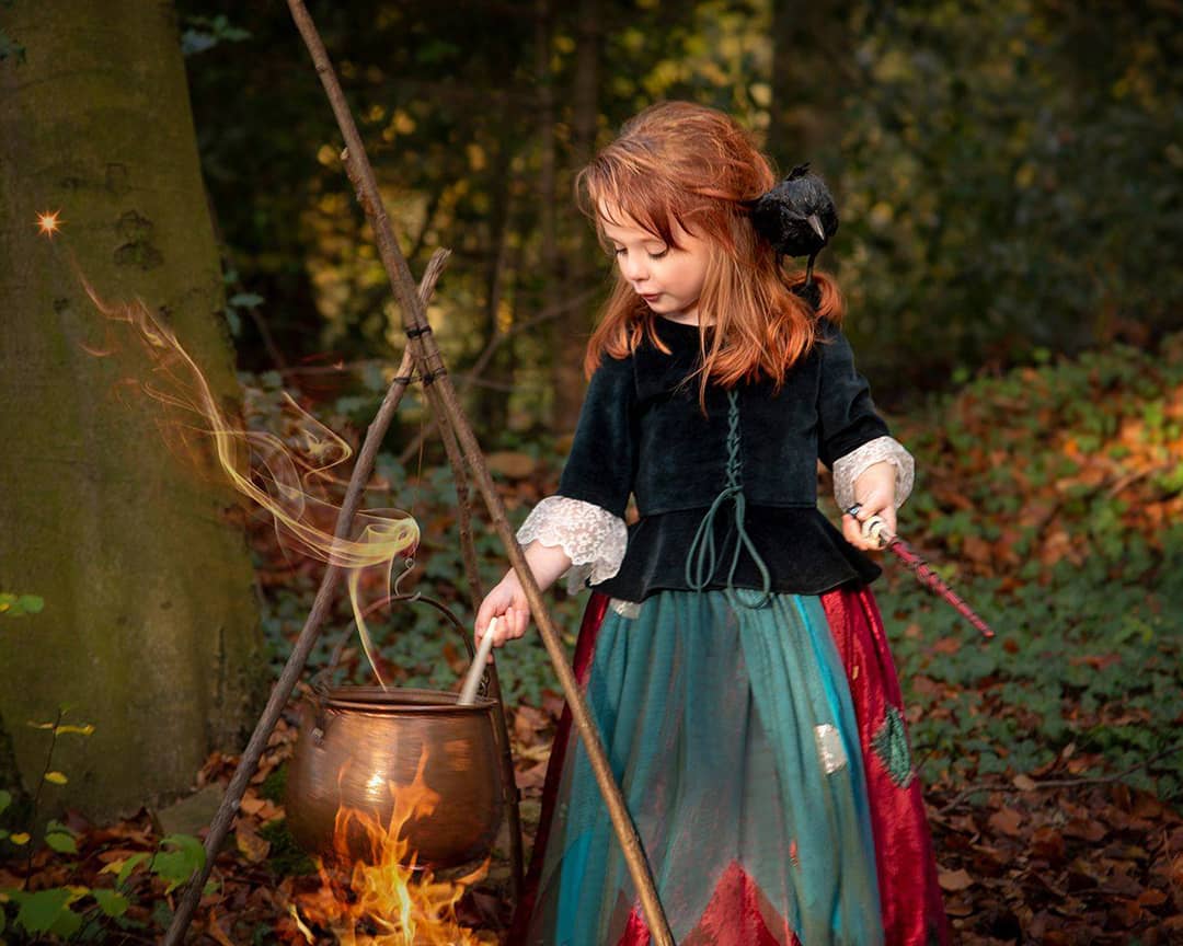 Ein kleines rothaariges Mädchen im kleine Hexe-Kleid rührt mit einem Zauberstab in einem rauchenden Hexenkessel über einem Lagerfeuer im Wald beim Märchenshooting in Wuppertal