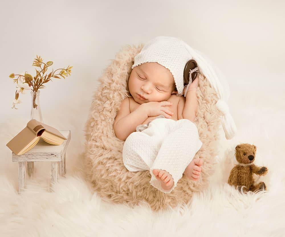 fotostudio-wuppertal-fuer-neugeborene-kreativ baby in einem winzigen beigen Sessel in cremetönen - neben sich ein Miniteddybär und ein Miniaturbuch auf einem Tisch beim Neugeborenenshooting in Wuppertal