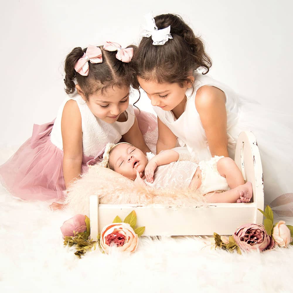 geschwisterliebe-beim-babyshooting-im-fotostudio-wuppertal 2 kleine maedchen mit dunklen Locken beugen sich lächelnd über ihre baby-schwester im Minibett bei einem Neugeborenenshooting in Wuppertal