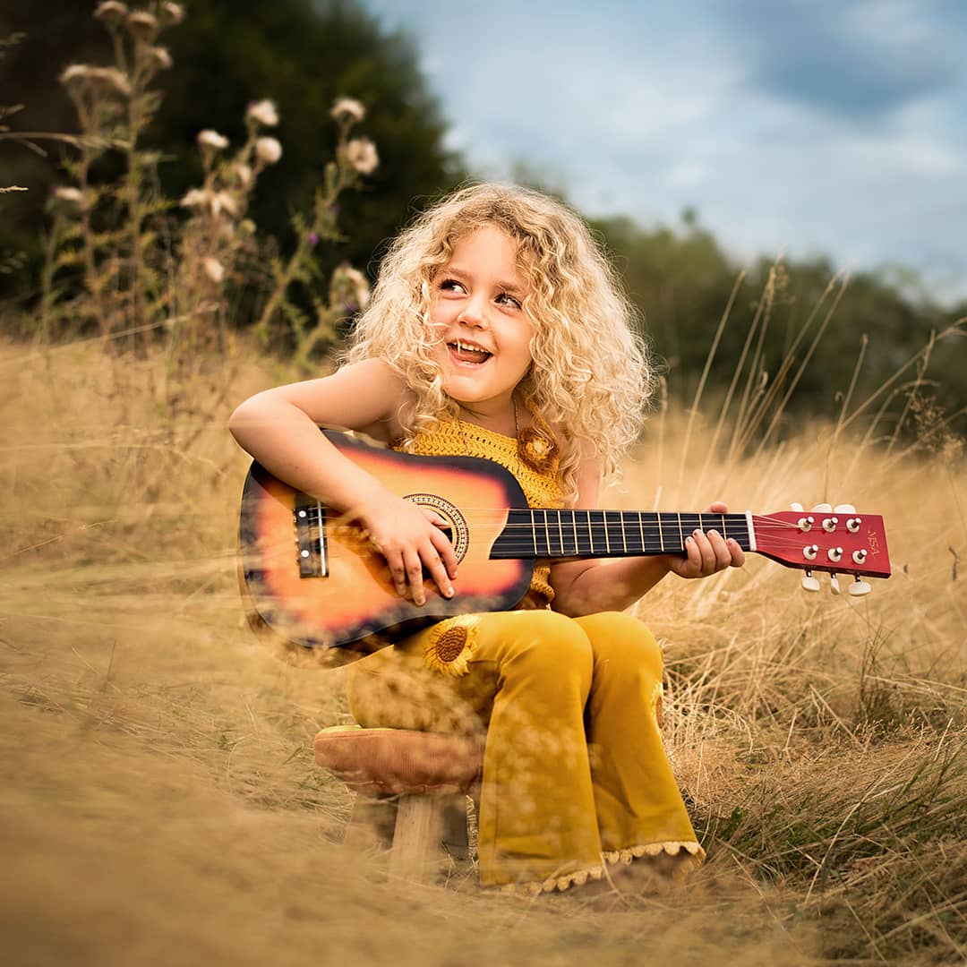 Ein herzhaft lachendes Kind sitzt mit einer Gitarre im Sommergras und strahlt voller Freude während eines echten und ungezwungenen Abenteuer-Fotoshootings in NRW.