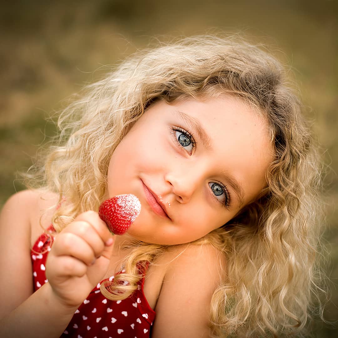 Blondes Mädchen mit wunderschönen blauen Augen blickt verträumt in die Kamera und hält eine gezuckerte Erdbeere beim professionellen Kinder-Porträtshooting in Wuppertal