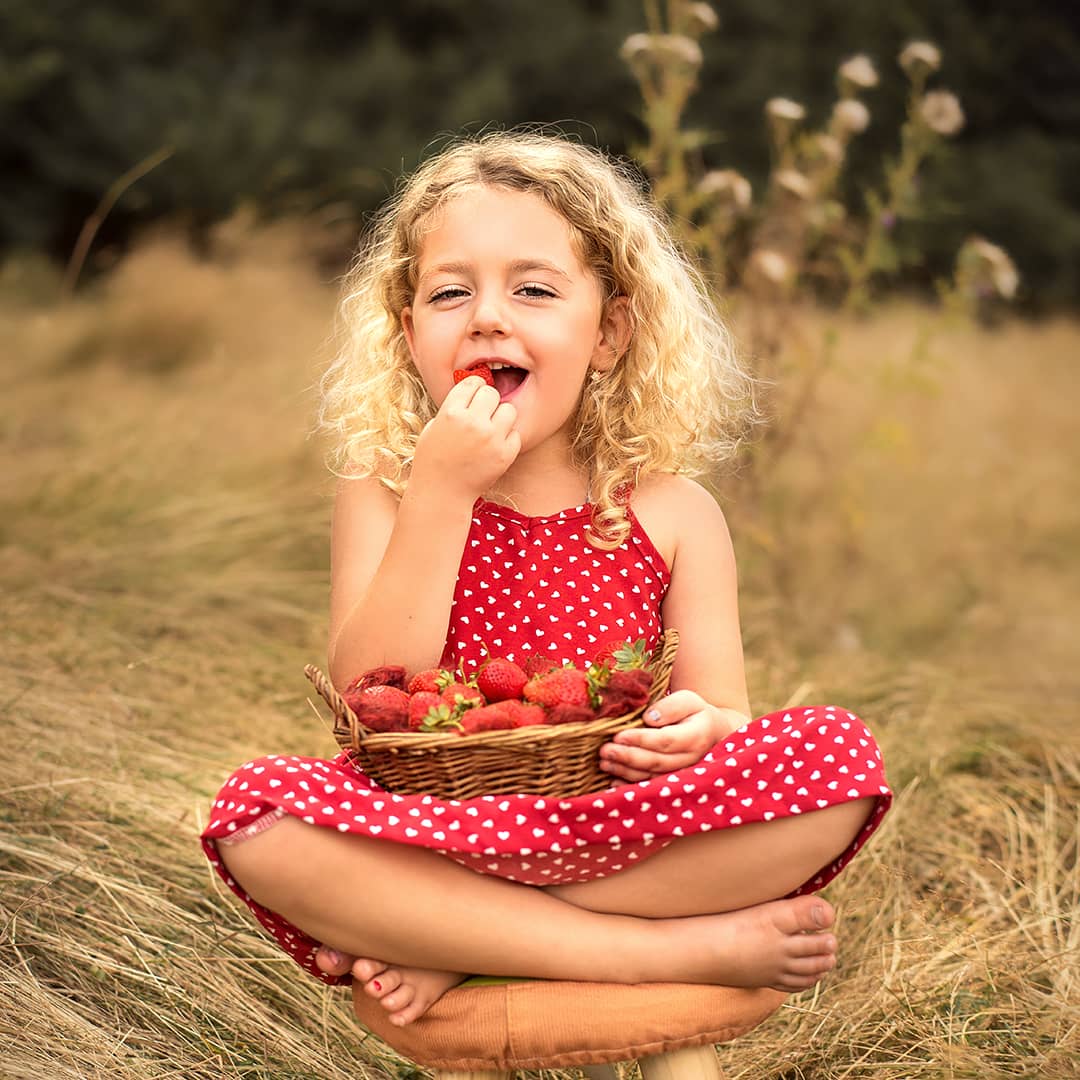 Mädchen im roten Sommerkleid mit einem Korb Erdbeeren im Schoß beißt genussvoll lächelnd in eine Erdbeere beim Sommer-Mini-Shooting für Kinder in Wuppertal