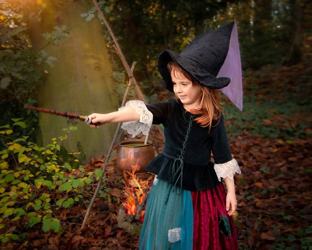 Ein kleines Mädchen mit spitzem Hexenhut streckt den Zauberstab aus, während im Hintergrund ein Lagerfeuer unter einem Hexenkessel brennt beim Märchenfotoshooting in NRW