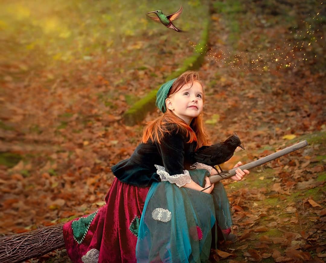 kleine Hexe Fotoshooting in Wuppertal: Ein kleines Mädchen sitzt mit einem Raben auf einem Hexenbesen im herbstlichen Laub und blickt staunend nach oben zu einem fliegenden Vogel den sie herbeigezaubert hat