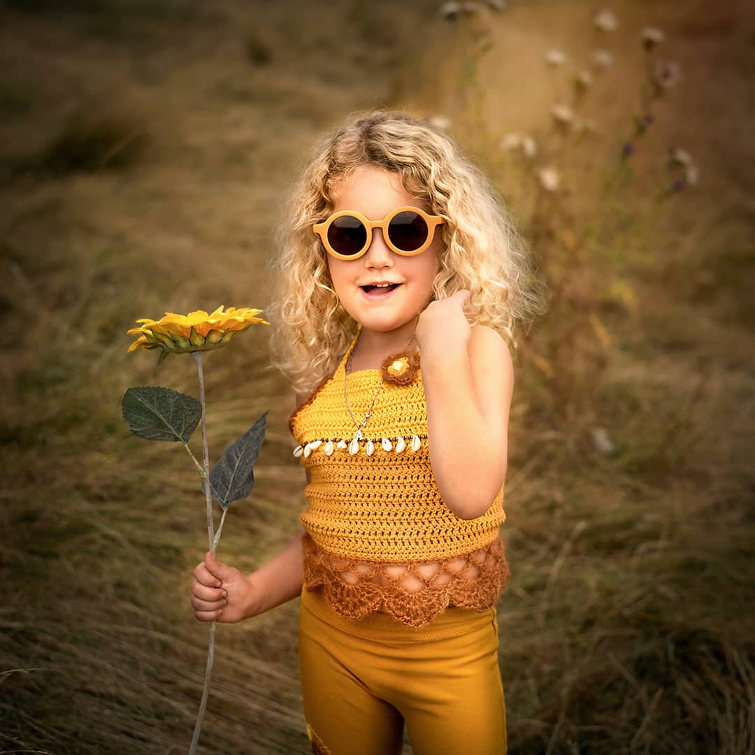 Künstlerische Kinderfotografie im Raum Wuppertal: Ein kleines Kind im gelben Boho-Outfit posiert witzig mit Sonnenbrille während eines erlebnisreichen Outdoor-Shoots