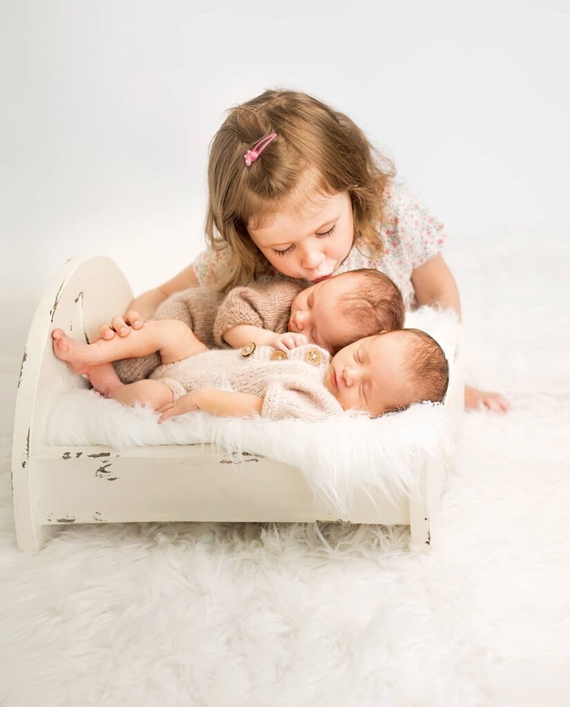 Ein kleines Mädchen küßt liebevoll die Zwillingsbabys im Bettchen vor ihr bei der Neugeborenen-Fotografie in Wuppertal