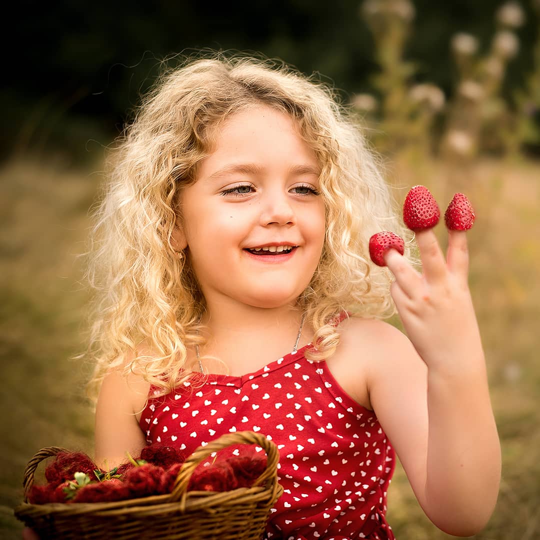Glückliches blondes Mädchen mit Erdbeeren auf den Fingern bei einem ungestellten Outdoor-Kinder-Fotoshooting in Wuppertal