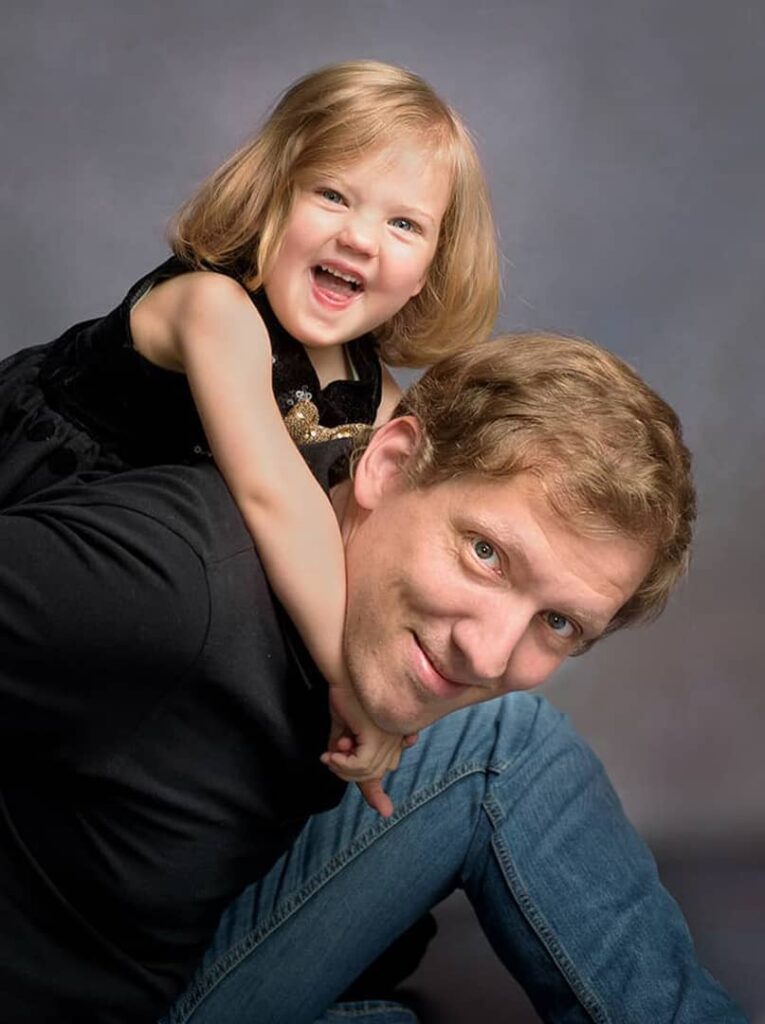 kleine blonde Tochter umarmt Vater lachend von hinten und schlingt die Arme um ihn, beim Fotoshooting in Wuppertal